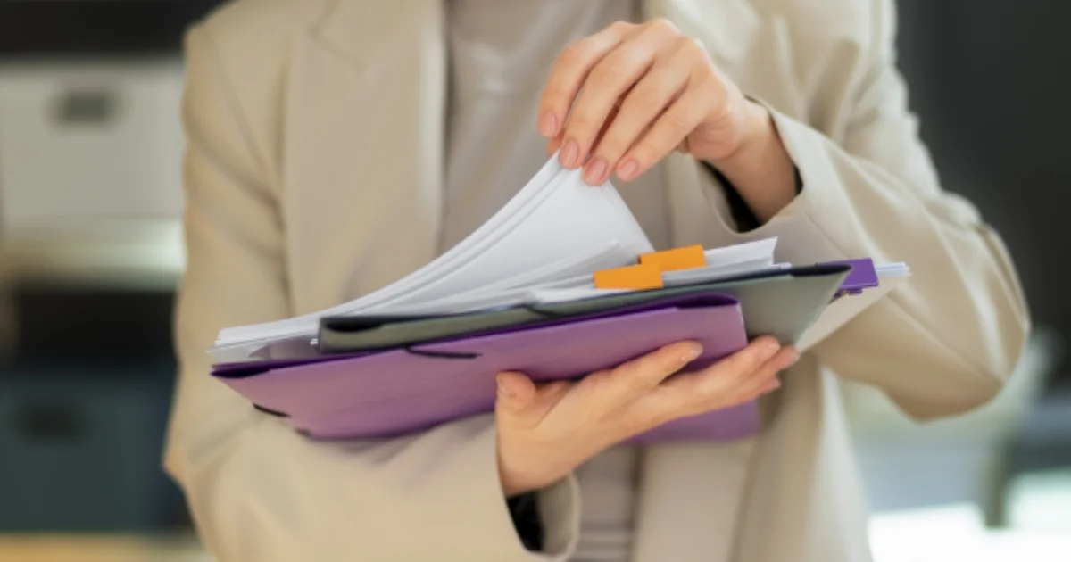 Person in a suit jacket flipping through a stack of folders and documents.